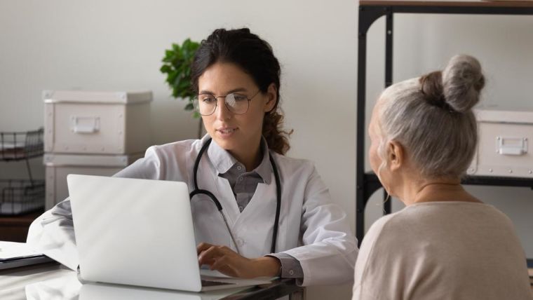 Serious young female doctor meeting with elderly patient in office, listening to woman health problems complaints, typing on laptop, keeping electronic database on computer. Geriatric medic care.